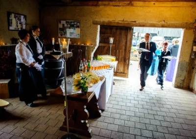 Ein Raum mit Kerzenlicht, Servicemitarbeiter am Tisch mit Getränken; Personen betreten den Raum, umgeben von rustikalem Ambiente.