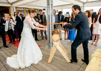 Das Brautpaar zersägt gemeinsam einen Baumstamm vor einer Gruppe von Gästen, die die Szene fotografieren.