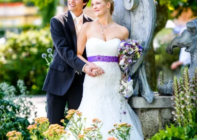 Ein Brautpaar steht lächelnd im Garten vor einem steinernen Denkmal; die Braut hält einen violetten Brautstrauß.
