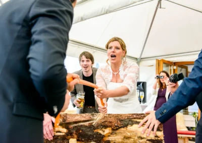 Braut im weißen Kleid sägt mit Bräutigam an einem Baumstamm. Gäste schauen zu, einige fotografieren. Fröhliche Stimmung.