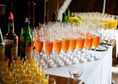 Sektgläser mit orangem Drink und Teelicht auf weiß gedecktem Tisch, umgeben von Flaschen und Orangensaftgläsern.