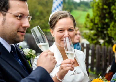 Ein Hochzeitspaar stößt mit Sektgläsern im Freien an, die Braut trägt ein weißes Kleid und lächelt in die Kamera.