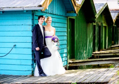 Ein Brautpaar lehnt lächelnd an einer türkisfarbenen Holzwand; die Braut hält einen lila Strauß, der Bräutigam trägt einen Anzug.