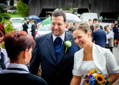 Brautpaar lächelnd im Regen, Bräutigam mit Anzug und Ansteckblume, Braut in Weiß mit Blumenstrauß, Gäste im Hintergrund.