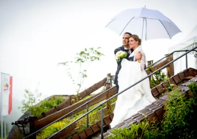 Ein Brautpaar unter einem weißen Regenschirm geht lächelnd eine Treppe herab, die Braut hält einen Blumenstrauß.