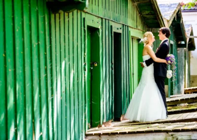 Ein Brautpaar lacht sich an, die Braut im weißen Kleid hält einen bunten Blumenstrauß, sie stehen vor grünen Holzhütten.