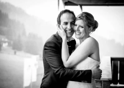 Ein lächelndes Brautpaar umarmt sich vor einer nebligen, bewaldeten Kulisse. Die Braut trägt ein Spitzenkleid.