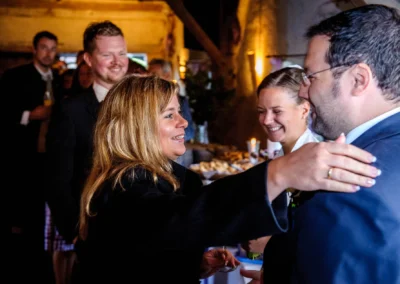 Eine blonde Frau gratuliert einem Mann im Anzug, im Hintergrund Gäste und ein Buffet, entspannte Atmosphäre.