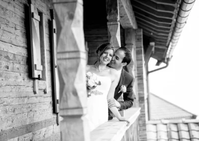 Ein Brautpaar lächelt auf einem Holzbalkon. Die Braut trägt ein schulterfreies Kleid und hält einen Brautstrauß.