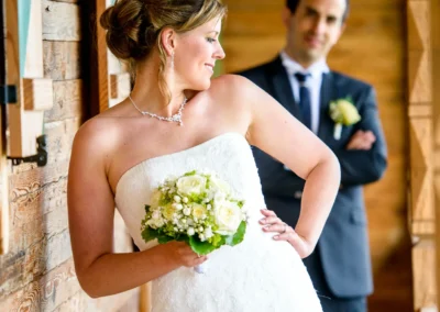 Die Braut in weißem Kleid hält einen Blumenstrauß, lächelt zur Seite; im Hintergrund der Bräutigam in dunklem Anzug.