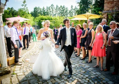 Das Brautpaar spaziert fröhlich Hand in Hand, umgeben von lächelnden Gästen in sommerlicher Kleidung. Hochzeitsreportage in Taufkirchen bei München.