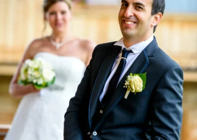 Ein Bräutigam in dunklem Anzug lächelt, im Hintergrund steht die lächelnde Braut mit Brautstrauß vor einem Holzgebäude.