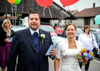 Das Brautpaar hält bunte Ballons, die Braut trägt einen Blumenstrauß. Gäste stehen im Hintergrund auf einem Parkplatz.