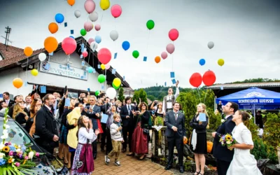 Eine Hochzeitsgesellschaft lässt bunte Luftballons steigen, das Brautpaar schaut lächelnd zu; Fotografiert von einem Hochzeitsfotografen in Moosach bei Grafing