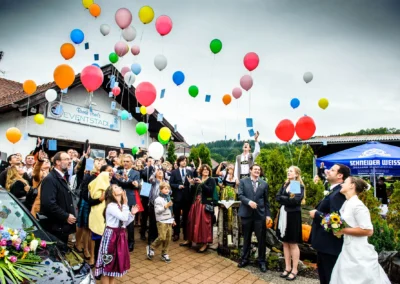 Eine Hochzeitsgesellschaft lässt bunte Luftballons steigen, das Brautpaar schaut lächelnd zu; Fotografiert von einem Hochzeitsfotografen in Moosach bei Grafing