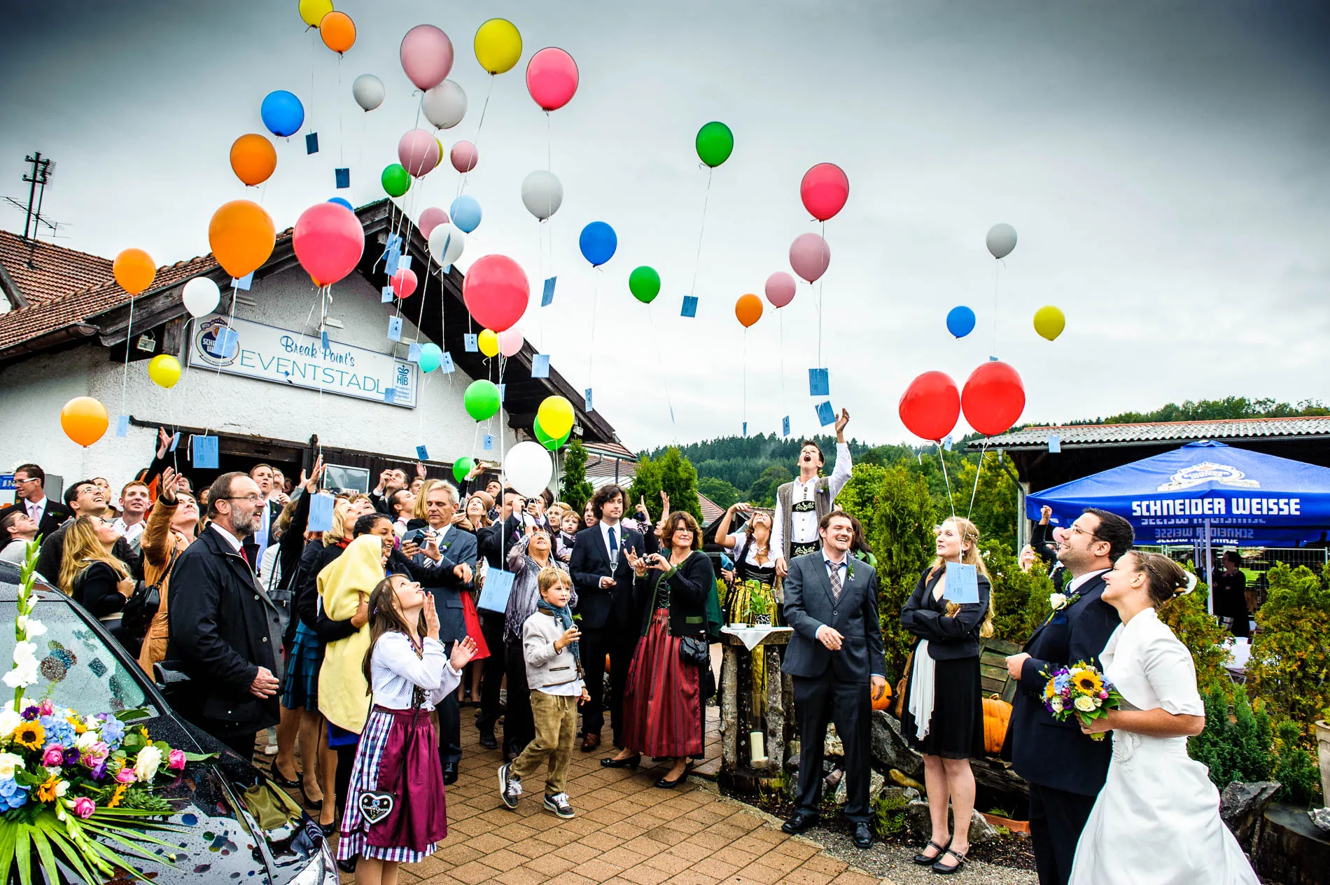 Eine Hochzeitsgesellschaft lässt bunte Luftballons steigen, das Brautpaar schaut lächelnd zu; Fotografiert von einem Hochzeitsfotografen in Moosach bei Grafing