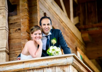 Das Brautpaar lächelt auf einem Holzbalkon. Die Braut hält einen weißen Blumenstrauß, der Bräutigam trägt einen Anzug.