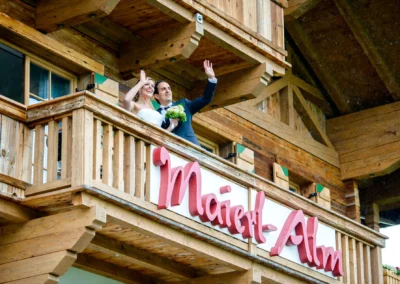 Brautpaar winkt fröhlich vom Balkon eines hölzernen Gebäudes. Hochzeitsreportage in Kirchberg bei Kitzbühel.