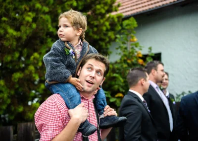 Ein Mann in kariertem Hemd trägt ein Kind auf den Schultern, beide schauen freudig. Im Hintergrund stehen mehrere Personen.