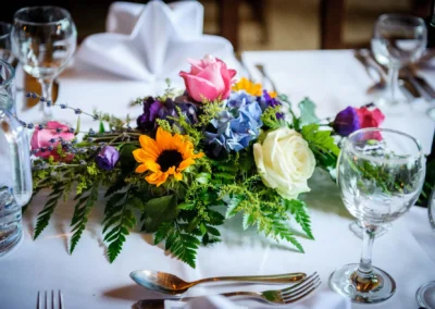 Bunter Blumenstrauß mit Sonnenblume, Rose und Hortensie auf weißer Tischdecke, umgeben von Gläsern und Besteck.
