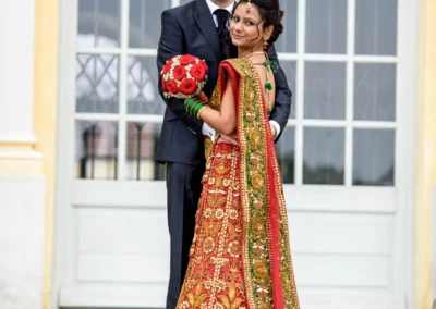 Brautpaar vor einer Tuer in Schloss Schleissheim. Die Braut traegt ein verziertes rotes Kleid und der Bräutigam einen Anzug.