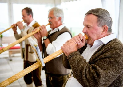 Drei Männer spielen Alphörner in traditioneller Kleidung in einem geschmückten Zelt.