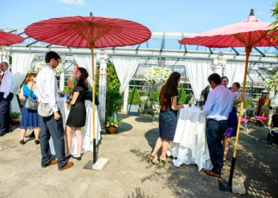 Gäste unter roten Sonnenschirmen plaudern an Stehtischen, umgeben von weißer Dekoration und Pflanzen im Freien.