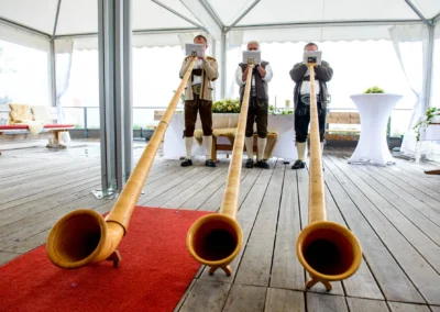 Drei Männer spielen Alphorn in traditioneller Kleidung auf einer Holzterrasse unter einem Zelt.