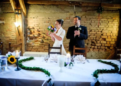 Braut mit Blumenstrauß und Bräutigam klatschen an einem festlich gedeckten Tisch vor roher Ziegelwand.