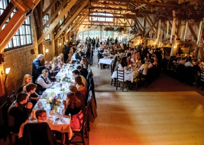 In einer festlichen, rustikalen Scheune sitzen Gäste an langen, weiß gedeckten Tafeln. Dezente Dekorationen und Kerzenlicht sorgen für eine warme Atmosphäre.