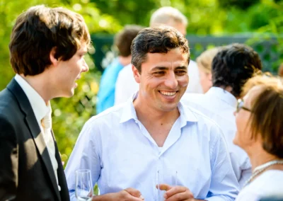 Zwei Männer und eine Frau unterhalten sich fröhlich im Freien. Sie halten Sektgläser und tragen elegante Kleidung.