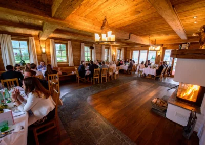 Gäste sitzen an gedeckten Tischen in einem rustikalen Raum. Ein Kamin brennt, warme Atmosphäre, Holzdekor.