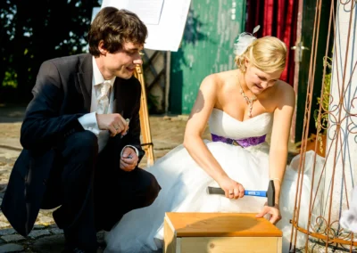 Das Brautpaar lächelt und hämmert gemeinsam auf eine Holzbox. Sie trägt ein weißes Kleid, er einen dunklen Anzug.