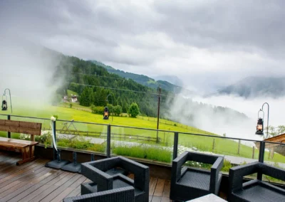 Terrasse mit Rattanmöbeln, Blumenstrauß am Geländer, Laternen. Nebliges Tal, grüne Hügel im Hintergrund.