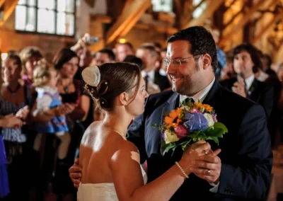 Ein Brautpaar tanzt lächelnd, die Braut hält einen bunten Blumenstrauß. Gäste im Hintergrund applaudieren.