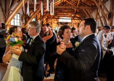Ein Brautpaar und weitere Gäste tanzen eng umarmt in einer festlich dekorierten Scheune. Die Braut hält einen Blumenstrauß.