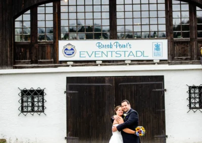 Das Brautpaar umarmt sich vor Eventstadl zur alten Säge in Moosach/ Grafing die Braut hält einen bunten Blumenstrauß.