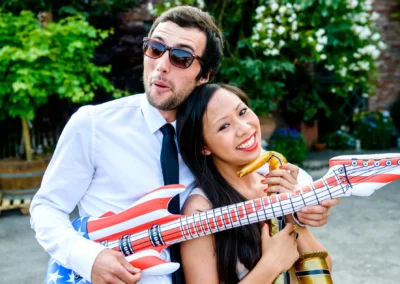 Zwei Personen lachen mit aufblasbaren Musikinstrumenten, Background aus grünen Pflanzen und Ziegelwand.