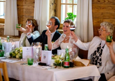 Eine fröhliche Hochzeitsgesellschaft stößt an einem gedeckten Tisch mit Weingläsern an, die Braut lächelt strahlend.