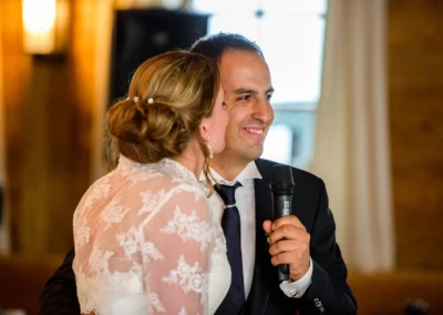 Ein Paar umarmt sich, er hält ein Mikrofon, sie trägt ein Spitzenkleid, beide lächeln. Hintergrund mit Holzdetails.