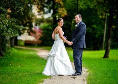 Ein Brautpaar steht auf einem Kiesweg im Park, Hand in Hand lächelnd, die Braut in weißem Kleid, der Bräutigam im Anzug.
