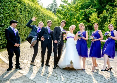 Eine fröhliche Hochzeitsgesellschaft posiert auf Kopfsteinpflaster, Braut in weiß, Bräutigam und Begleiter in lila Akzenten.