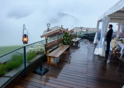 Eine überdachte Terrasse im Regen mit Lampen, Bänken und einem Mann im Anzug, Berge im Nebel im Hintergrund.