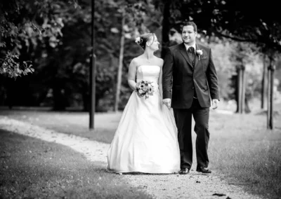 Brautpaar spaziert auf einem Weg im Park, die Braut trägt ein langes Kleid, der Bräutigam einen Anzug. Sie hält einen Blumenstrauß.