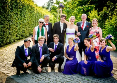 Eine fröhliche Hochzeitsgruppe posiert draußen, Braut in weißem Kleid, Gäste in lila und schwarzen Anzügen, umrahmt von grünem Laub.