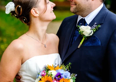 Das Brautpaar lächelt sich an, sie hält einen bunten Blumenstrauß, er trägt einen dunklen Anzug mit Blumenanstecker.