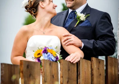 Ein Brautpaar steht lächelnd hinter einem hölzernen Zaun. Sie trägt ein weißes Kleid und einen bunten Blumenstrauß.