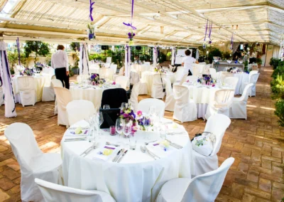 Ein festlich gedeckter Raum mit weißen Tischdecken, lila Blumendekorationen und Gästen in eleganter Kleidung.