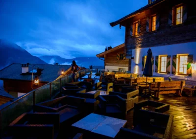 Eine Person steht auf einer Terrasse bei Dämmerung; Lichter leuchten, rustikale Holzmöbel und Berge im Hintergrund.