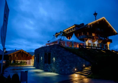 Beleuchtetes Haus mit großen, spitzen weißen Zeltdächern bei Dämmerung, beleuchtete Steinmauer und Treppe im Vordergrund.